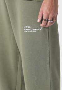 Housut oliivinvihreästä puuvillasekoitteesta, jossa on valkoisella painettu "IRASUTO STUDIOS" -logo, rento malli ja sivutaskut.