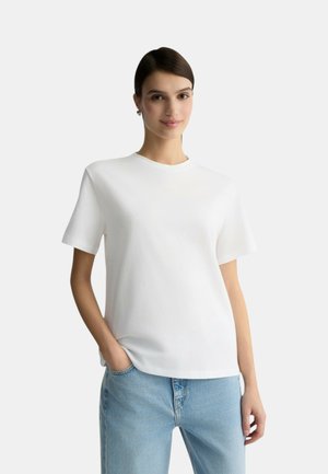 T-shirt bianca in cotone con collo rotondo e maniche corte, abbinata a jeans azzurri chiari. Design semplice, texture morbida e vestibilità rilassata.
