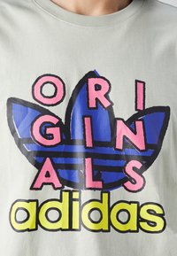 Světle zelené bavlněné tričko s velkým černým a modrým trojlístkovým logem, s růžovým textem "ORIGINALS" nahoře a žlutým "adidas" dole.