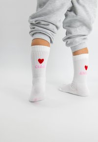 Weiße gerippte Socken mit roten Herzmotiven und den Worten "CIAD" und "AMOR" in pinker Schrift. An Füßen getragen, kombiniert mit grauen Hosen.