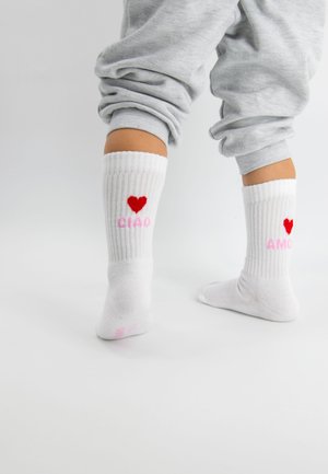 Weiße gerippte Socken mit roten Herzmotiven und den Worten "CIAD" und "AMOR" in pinker Schrift. An Füßen getragen, kombiniert mit grauen Hosen.