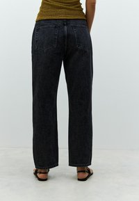 Schwarze gerade Jeans mit verblasster Textur, fünf Taschen und einem sauberen Saum, getragen mit schwarzen Sandalen vor einem neutralen Hintergrund.