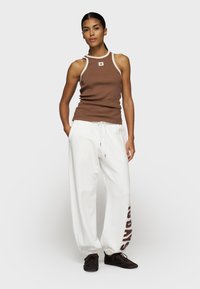 Braunes geripptes Tanktop mit weißem Saum und Logotag, kombiniert mit weißen Jogginghosen mit braunem Text. Schwarze Sneakers runden das Outfit ab.