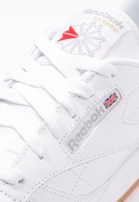 Weiße Reebok-Sneaker mit einem Logo in Rot- und Grautönen, strukturiertem Leder, flachen weißen Schnürsenkeln und einem kleinen Union-Jack-Detail.