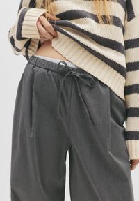 Grå afslappede bukser med snor i taljen, plisseret for og glat tekstur, parret med en cremet og marineblå stribet strikpullover.