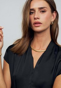 Svart blus med kort ärm och V-ringning. Bärs med ett halsband i guldmetall och ett matchande bangle-armband. Slät textur.