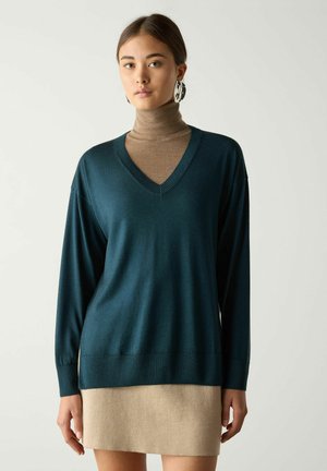 Kvinde iført teal V-hals sweater over brun rullekrave og beige nederdel, med store sorte og hvide øreringe, stående mod en ensfarvet baggrund.