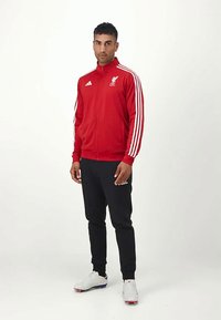 Chaqueta deportiva roja con rayas blancas, logo de Liverpool. Pantalones negros con logo de Adidas, acompañados de zapatillas deportivas blancas. Fondo liso.