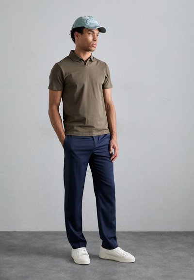 Brown polo shirt, navy trousers, and white sneakers. Light blue cap with embroidered detail. Standing against a neutral background.
