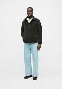 Hnedá košeľová bunda z kordovanej látky s gombíkmi vpredu a špicatým golierom, kombinovaná so svetlomodrými širokými džínsami a čiernymi topánkami.