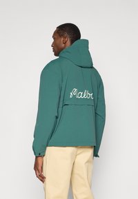 Ελαφρύ τζάκετ σε τιρκουάζ χρώμα με κουκούλα, διαθέτοντας κλείσιμο με κορδόνι και κρεμ κεντημένη επιγραφή "Malba" στην πλάτη. Απλή υφή, χωρίς ορατά σχέδια.