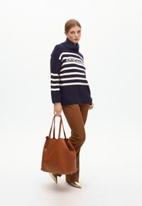 Námořnicky pruhovaný svetr s krémovými pruhy a logem, hnědé kalhoty a prošívaná hnědá kožená taška. Modelka má na sobě zlaté lodičky se špičatou špičkou.