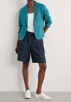 Persona con una giacca verde acqua sopra una maglia bianca, pantaloncini scuri al ginocchio e sneakers bianche, in piedi contro un muro semplice.
