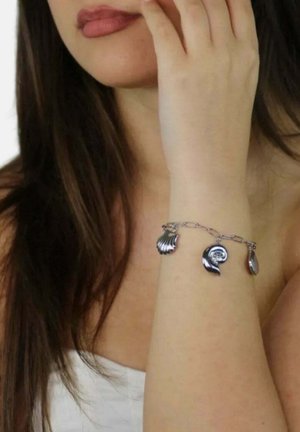 Mujer con cabello largo y oscuro que lleva una pulsera de plata con dijes de conchas marinas y espirales en la muñeca, con la mano cerca de su rostro.