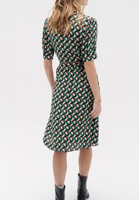 Groene jurk met geometrische patronen in rood, wit en zwart. Korte mouwen, gestrikte taille en vloeiende silhoeet. Gemaakt met zwarte enkellaarsjes.