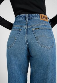 Blaue Denim-Jeans mit hoher Taille und einem hellbraunen Lederlabel, ausgestattet mit Gesäßtaschen, die von gelben Nähten umrandet sind, und einem geraden Schnitt.