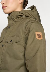 Geacă cu glugă de culoare verde măsliniu, cu un finisaj texturat, având două buzunare frontale și o etichetă din piele cu un logo pe mânecă.