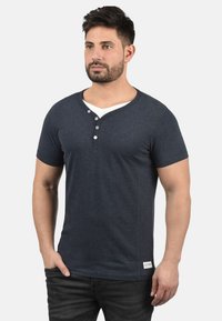 Jūras spēku īso piedurkņu henley T-krekls, izgatavots no mīksta auduma, ar V veida kaklu un trim pogām, kā arī mazu logotipa uzšuvi apakšā.