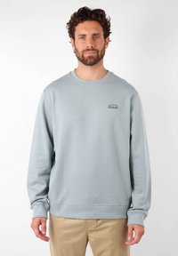 Sweatshirt gris clair en tissu doux, avec un col rond, des manches longues et un petit logo vert sur la poitrine.
