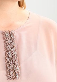 Rosa blus i genomskinligt tyg, med en central utsmyckning av samlade pärlor och paljetter. Mjuk textur och enkel design.