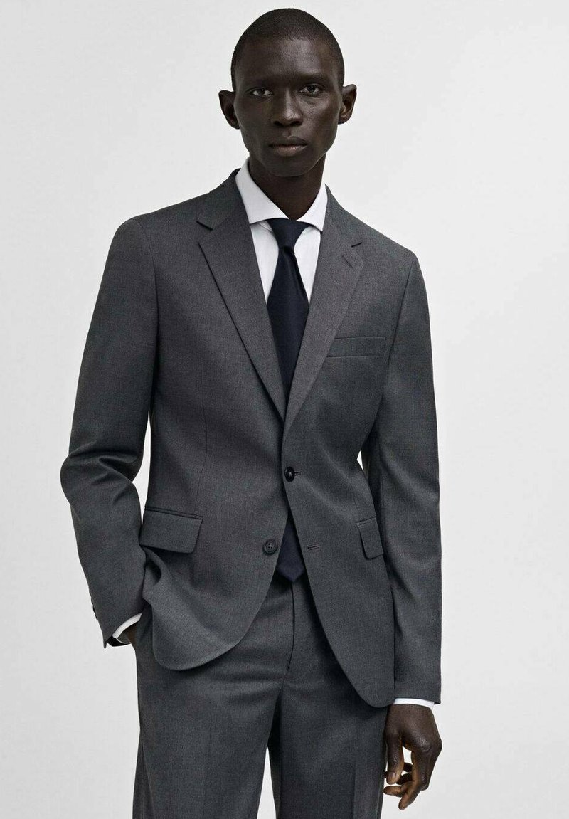 Completo grigio con chiusura a due bottoni, revers a punta, taschino sul petto. Camicia bianca e cravatta nera completano il look formale.