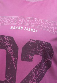 Fioletowa bawełniana koszulka z wyraźnym czarnym nadrukiem "02" oraz napisem "TRUE RELIGION BRAND JEANS®", o teksturowanym wykończeniu.