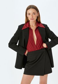 Blazer nero con rever affilati sopra una camicia bordeaux, abbinato a una gonna avvolgente nera a vita alta. Tessuto morbido, vestibilità su misura.