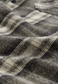 Tissu en flanelle doux avec un motif à carreaux noir, gris et blanc, montrant des détails tissés texturés et un bouton noir visible.
