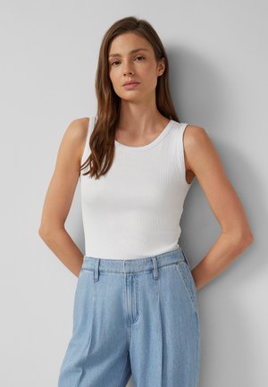 Kobieta z długimi brązowymi włosami, ubrana w biały bezrękawnik z prążkowanego materiału i jasnoniebieskie dżinsy z wysokim stanem, stojąca przy szarej ścianie.