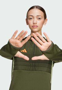 Dark green athletic jacket with a zippered front, featuring a padded chest area, orange logo accents, and textured sleeves. Hands positioned outward.