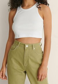 Weißes, geripptes Cropped Top mit hohem Kragen, kombiniert mit hochgeschnittenen olivgrünen Jeans, die mit Metallknöpfen und Vordertaschen versehen sind.