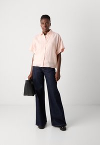 Camicetta a maniche corte in lino rosa chiaro, con bottoni davanti, abbinata a pantaloni svasati in denim scuro, tiene in mano una borsa in pelle nera. Design semplice e pulito.