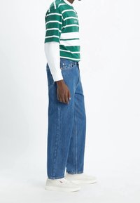 Hombre vistiendo una camisa de rayas verdes y blancas de manga corta sobre una camiseta blanca de manga larga, jeans azules y zapatillas blancas, de pie de lado.