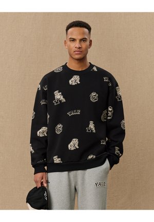 Sort sweatshirt med Yale-tema grafik i beige, inklusive bjørne og logoer. Har ribbet halsudskæring og manchetter. Matcher med grå sweatpants.