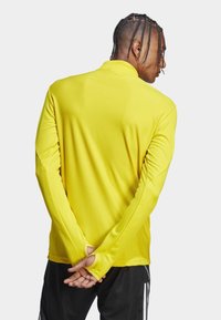 T-shirt de sport à manches longues jaune avec tissu texturé, col montant et ouvertures pour les pouces, associé à un pantalon noir avec des rayures blanches.