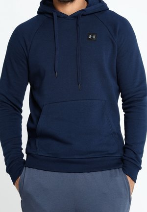 Mann trägt einen marineblauen Under Armour Hoodie mit Fronttasche und Kordelzügen, Hände teilweise in den Taschen, vor einfarbigem Hintergrund.
