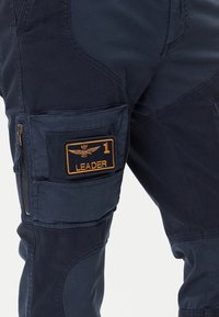 Pantaloni cargo in tessuto blu navy scuro con cuciture visibili, una tasca laterale con un'etichetta arancione con la scritta "LEADER" e un emblema alato, e chiusura con zip.