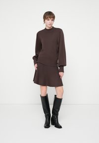 Kvinde står iført et brunt sæt med langermet sweater og nederdel, parret med sorte knæhøje støvler foran en ensfarvet hvid baggrund.