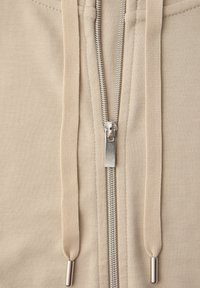 Tela beige con cremallera metálica plateada y dos cordones beige con puntas metálicas a cada lado, vista de cerca.