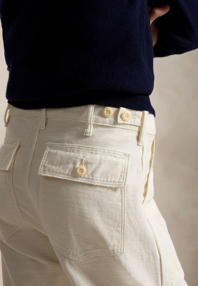 Persoană care poartă pantaloni bej deschis cu buzunare laterale și nasturi, asortați cu un top cu mânecă lungă bleumarin închis, arătat de la talie în jos.