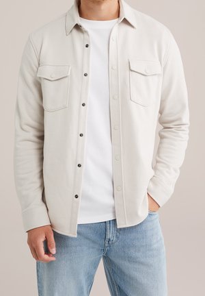 Mand iført en off-white skjorte med knapper og brystlommer over en hvid t-shirt og lyseblå jeans, med den ene hånd i lommen.