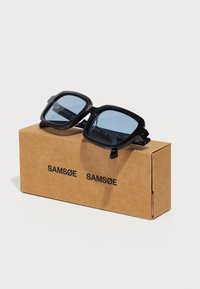 Ochelari de soare rectangulari negri cu lentile nuanțate de albastru, așezați pe o cutie de carton maro etichetată "SAM SøE". Grosime moderată și finisaj lucios.