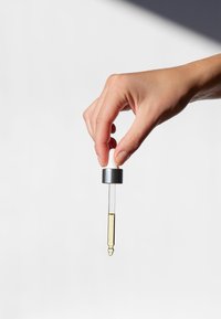 Held in een hand tegen een witte achtergrond is een heldere glazen pipetfles met een zilveren metalen dop, gevuld met een lichtgele vloeistof.