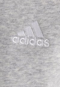 Šedá vřesová tkanina s jemnou strukturou je doplněna vyšitým bílým logem Adidas, které se skládá ze tří pruhů nad názvem značky.