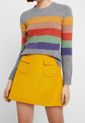 Femme portant un pull gris avec des rayures horizontales multicolores et une jupe jaune vif avec deux poches avant boutonnées.