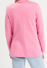 Veste rose à la texture lisse, au design ajusté, avec des manches longues. Le dos présente une couture centrale et des poches latérales. Associée à un jean bleu clair.