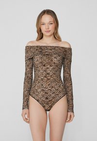 Mladá žena stojí v leopardím bodýčku s dlouhým rukávem a bez ramínek, čelí vpřed na jednoduchém světlém pozadí.