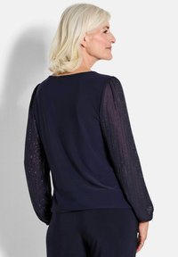 Blusa navy con maniche trasparenti e testurizzate adornate da linee verticali di piccole strass. Collo rotondo classico e silhouette aderente.