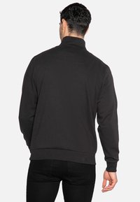 Giacca nera con zip, colletto alto, polsini e fondo a costine, realizzata in un materiale simile al jersey. Presenta cuciture minime e senza loghi visibili.