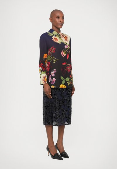 Μακρυμάνικη ανθισμένη μπλούζα σε σκούρο ύφασμα, συνδυασμένη με μια μαύρη μίνι φούστα με παγιέτες. Μαύρα ψηλά τακούνια σε σχήμα αιχμής ολοκληρώνουν το σύνολο.